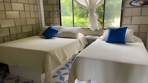 Deux lits simples avec du linge de lit blanc et des oreillers bleus, un sol carrelé et une fenêtre donnant sur un paysage verdoyant.