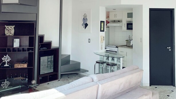 A modern living room with a sofa, a staircase, a kitchen area, and a door. - Entire Apartment - Duplex in Panamby (Vila Andrade São Paulo)