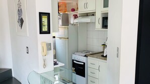 A compact kitchen with white cabinets, a microwave, and a small oven. There is a glass bar with two stools in the foreground. - Entire Apartment - Duplex in Panamby (Vila Andrade São Paulo)