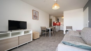 Een compact leefruimte met een flatscreen televisie op een witte tv-standaard, een zithoek met een tafel en stoelen, een kitchenette op de achtergrond, en een gestreept bankje.