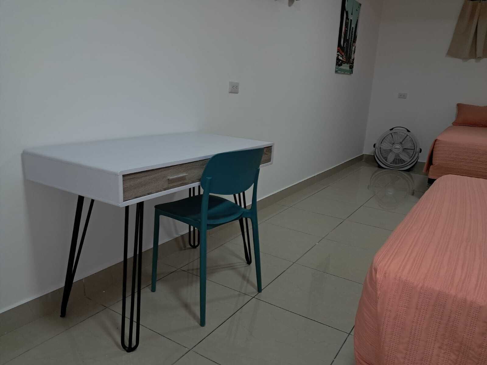 Een kleine, schone kamer met een wit bureau, een blauwgroene stoel en een bed met een roze sprei.