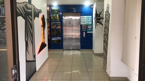 Un pasillo con un mural de una persona vestida de bata, un panel de control digital para el ascensor y un reloj en la pared.