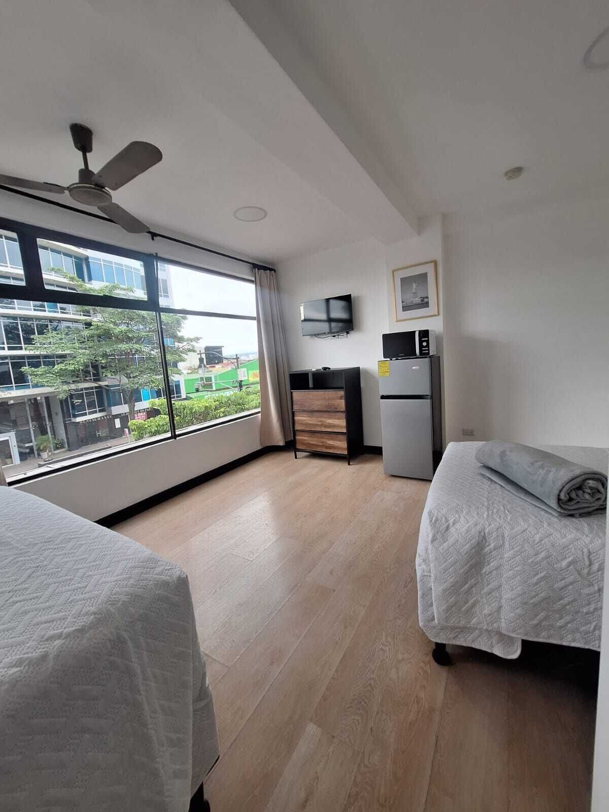 Une chambre d’hôtel moderne dotée d’une grande fenêtre, d’un ventilateur de plafond, d’un téléviseur à écran plat, d’un four à micro-ondes et d’un petit réfrigérateur.