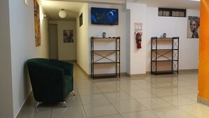 Un pasillo con un sillón verde, dos estanterías metálicas, un extintor y un televisor montado en la pared.