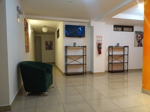 A hallway with a green armchair, two metal shelving units, a fire extinguisher, and a television mounted on the wall. - Apartahotel del Paseo (San José)