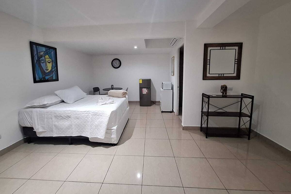 Ein Hotelzimmer mit Bett, kleinem Tisch mit Stühlen, Kühlschrank, einer Uhr und einem Spiegel an der Wand.