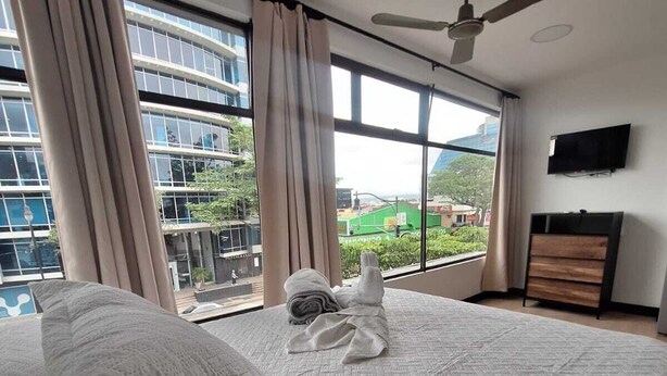 Un dormitorio con una cama grande, un ventilador de techo, un televisor en la pared y vistas de un paisaje urbano a través de amplios ventanales.
