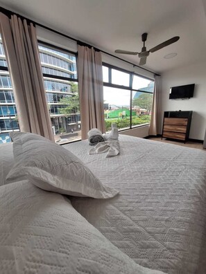 Un dormitorio con una cama grande, un ventilador de techo, un televisor en la pared y vistas de un paisaje urbano a través de amplios ventanales.