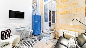 Een compacte leefruimte met een zwart-wit vloertegelpatroon, een blauwe deur, een witte radiator, een flatscreen-tv en een kleine ronde tafel.
