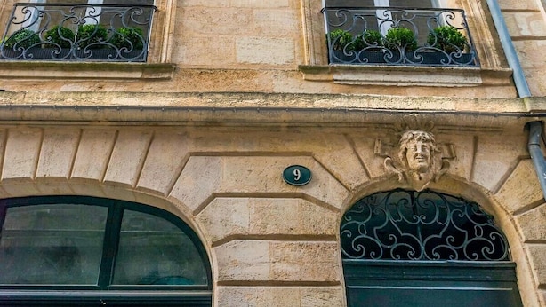 Een gebouw met drie ramen, elk met een balkon van smeedijzer en begroeiing. Het gebouw heeft een stenen gevel met een grote, rondboogvormige entree en een deur.