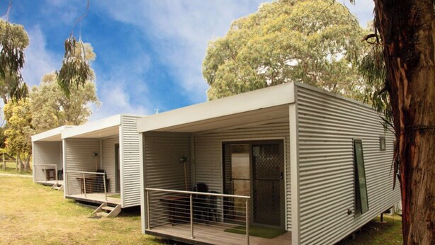 Une rangée de maisons mobiles modernes, de plain-pied, dotées de grandes fenêtres et d’un toit plat, entourées d’arbres.