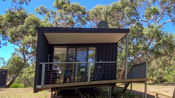 A small, dark-colored cabin with a balcony, surrounded by trees.