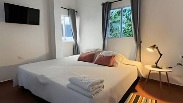 A modern hotel room with a large bed, a flat-screen TV mounted on the wall, a small bedside table with a lamp, and a window with curtains.