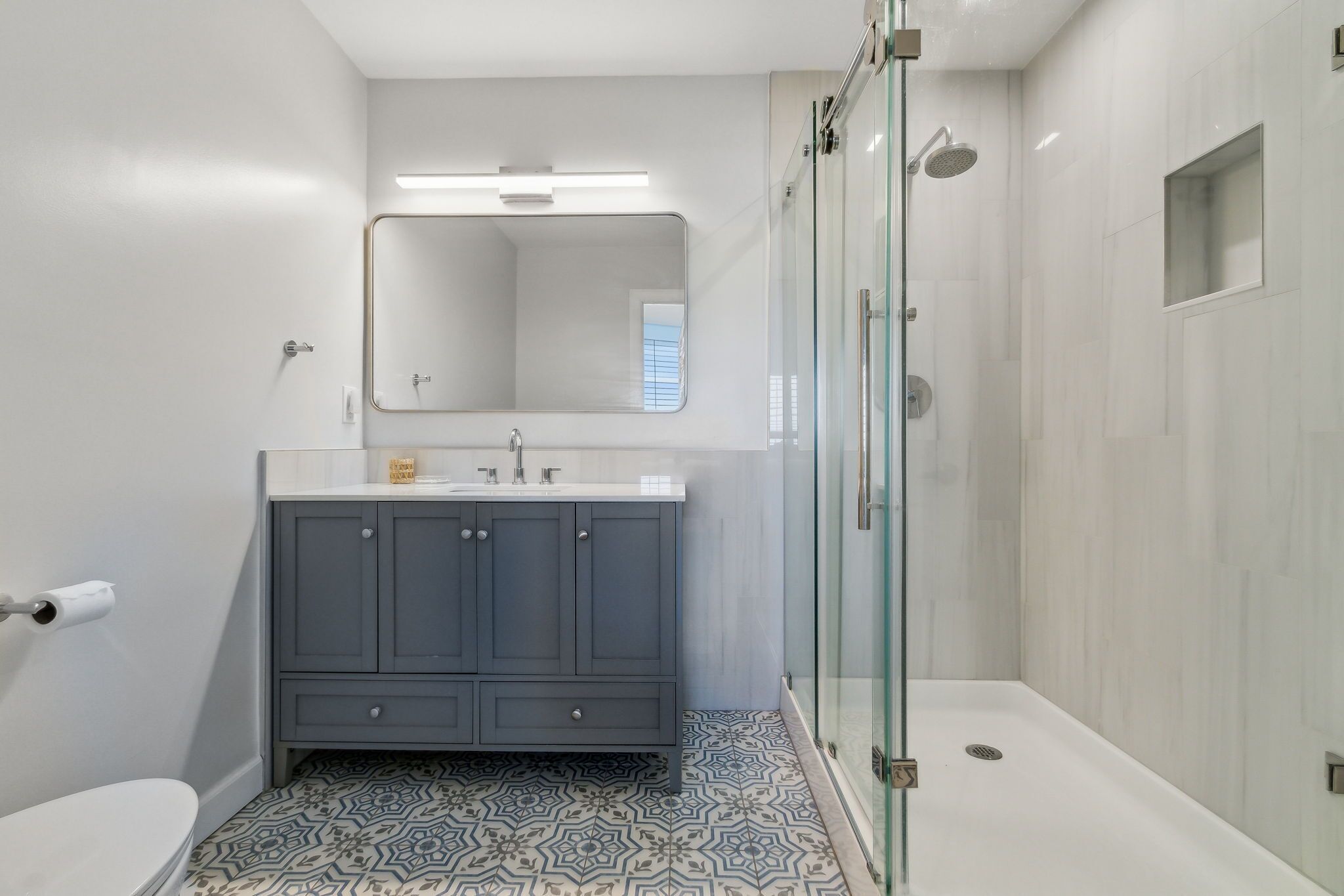 Un baño moderno con un inodoro blanco, un tocador gris con espejo y una zona de ducha con mampara de vidrio.