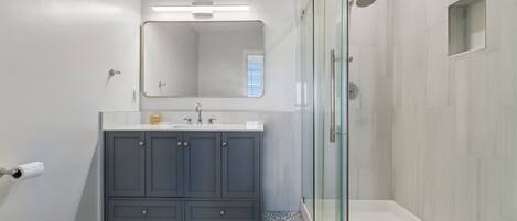 Un baño moderno con un inodoro blanco, un tocador gris con espejo y una zona de ducha con mampara de vidrio.