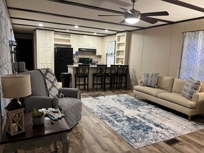 A modern living room with a sofa, a coffee table with decorative items, a rug, and a kitchen area with a refrigerator, microwave, and dining chairs. - Cozy 2-BR MH in wonderful country setting. Short drive to Industrial Plants (Chunchula)