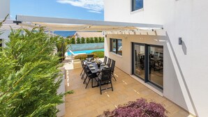 En moderne udendørs terrasse med spisebord og stole, en pool i baggrunden og en hvid bygning med store vinduer.