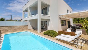 Et moderne hus med to etager, en swimmingpool, udendørs møbler og en overdækket terrasse.