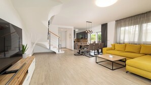 En moderne stue med en gul sofa, et træsofabord og et fjernsyn på et træstel. Der er en trappe til en etage ovenpå, og et spiseområde med stole og et bord.