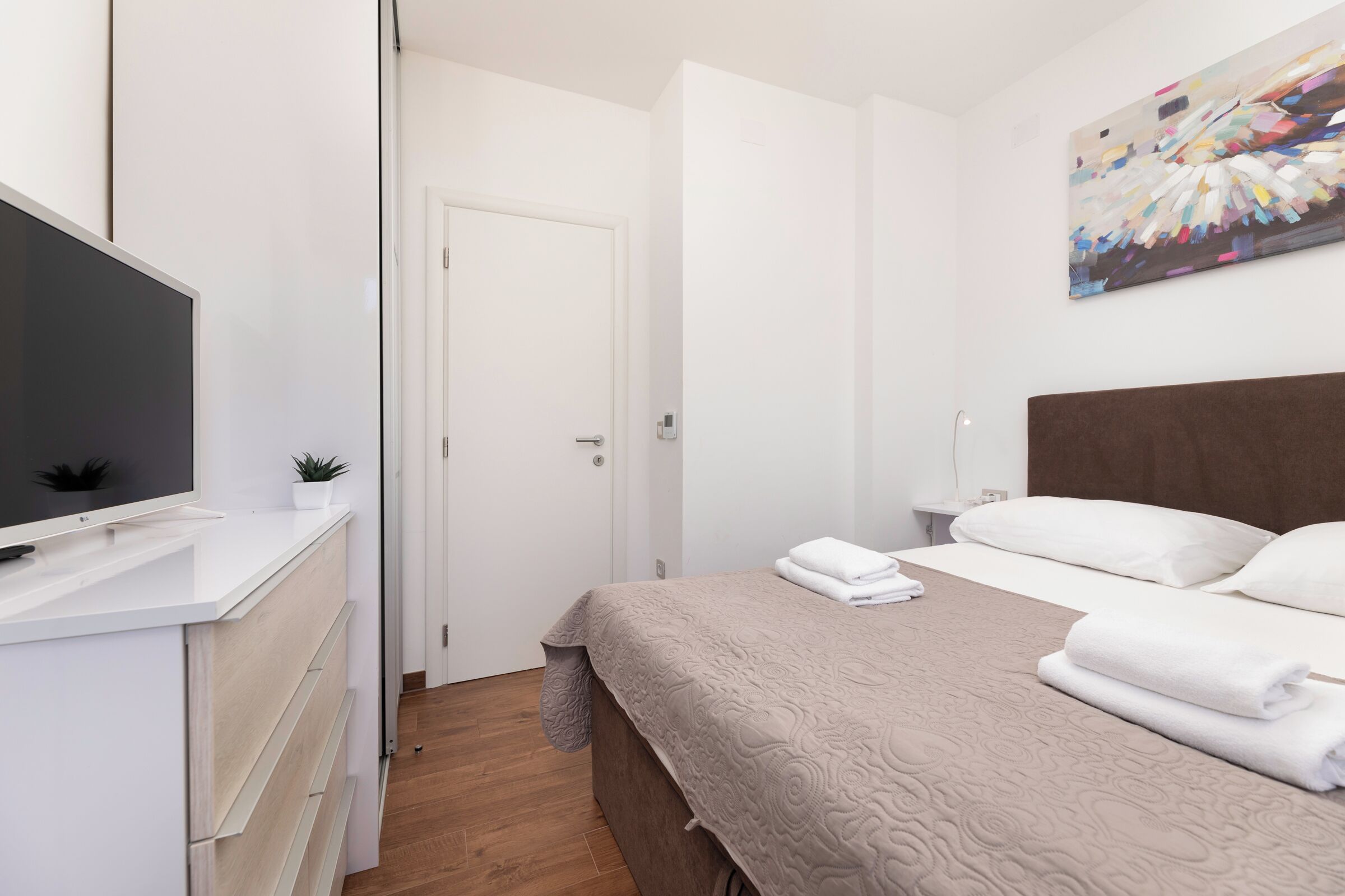 A modern hotel room with a bed, a TV, a painting, and a small plant.