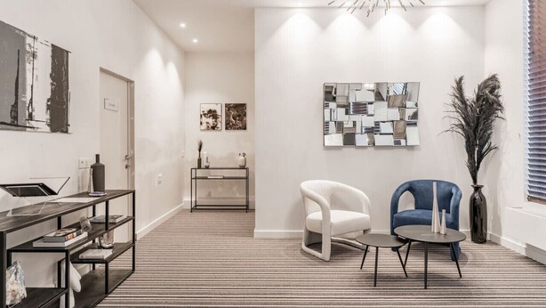 A modern living room with a black and white striped carpet, a white armchair, a blue armchair, a small round table, and a black console table with shelves. - Elegant room with private bathroom near city center (Brugge)