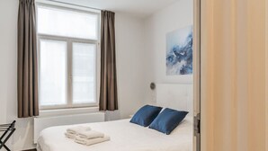 Une chambre à coucher avec un lit, deux oreillers, une fenêtre avec des rideaux, un radiateur et un tableau accroché au mur.