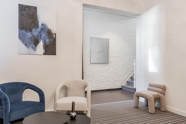 A modern room with a blue chair, a beige armchair, and a small stool. There is a painting on the wall and a staircase in the background.