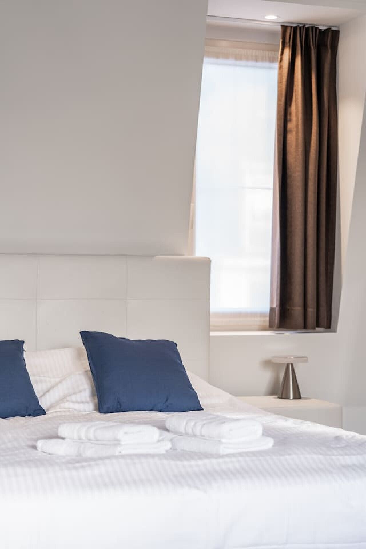 Un lit bien fait, avec du linge de lit blanc et des coussins bleus, une table de chevet avec une lampe et une fenêtre ornée de rideaux bruns.