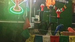Um quarto com um letreiro de neon, um narguilé e placas indicando um banheiro e um bar.