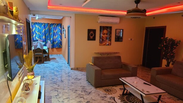 Una sala de estar con un sofá, sillones, una mesa de centro y un televisor. Hay una alfombra estampada en el suelo y un ventilador de techo.