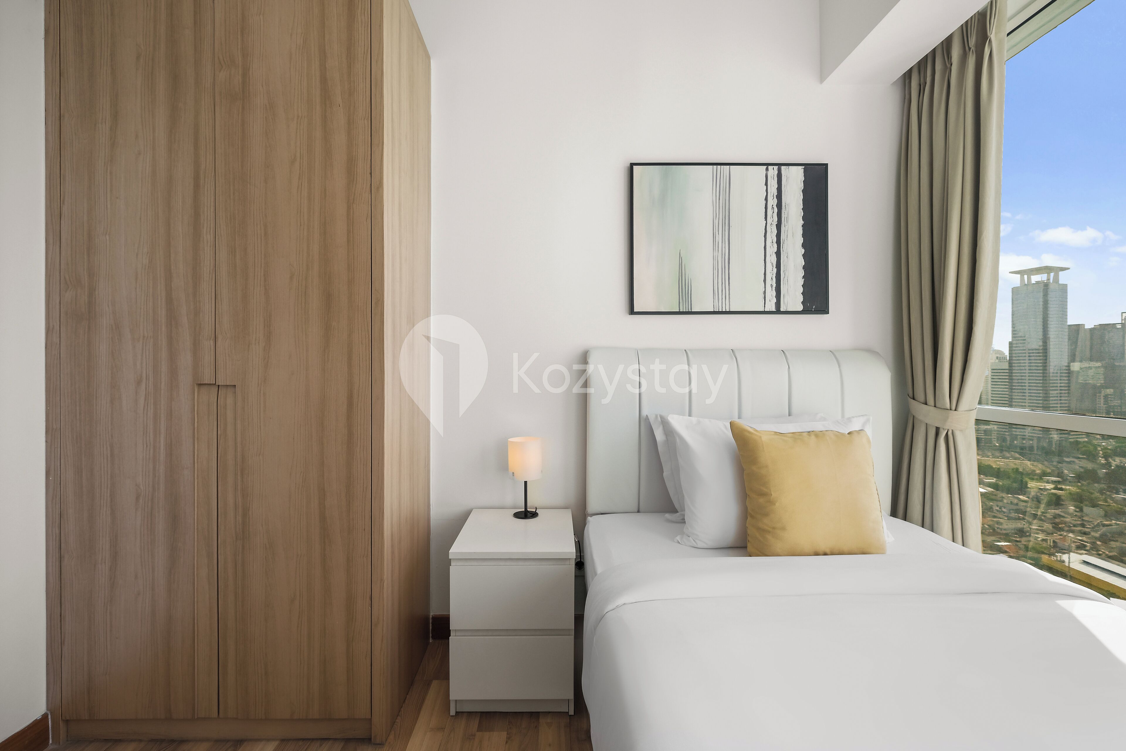 A modern hotel room with a large bed, a nightstand, a lamp, a city view, and a wooden wardrobe.