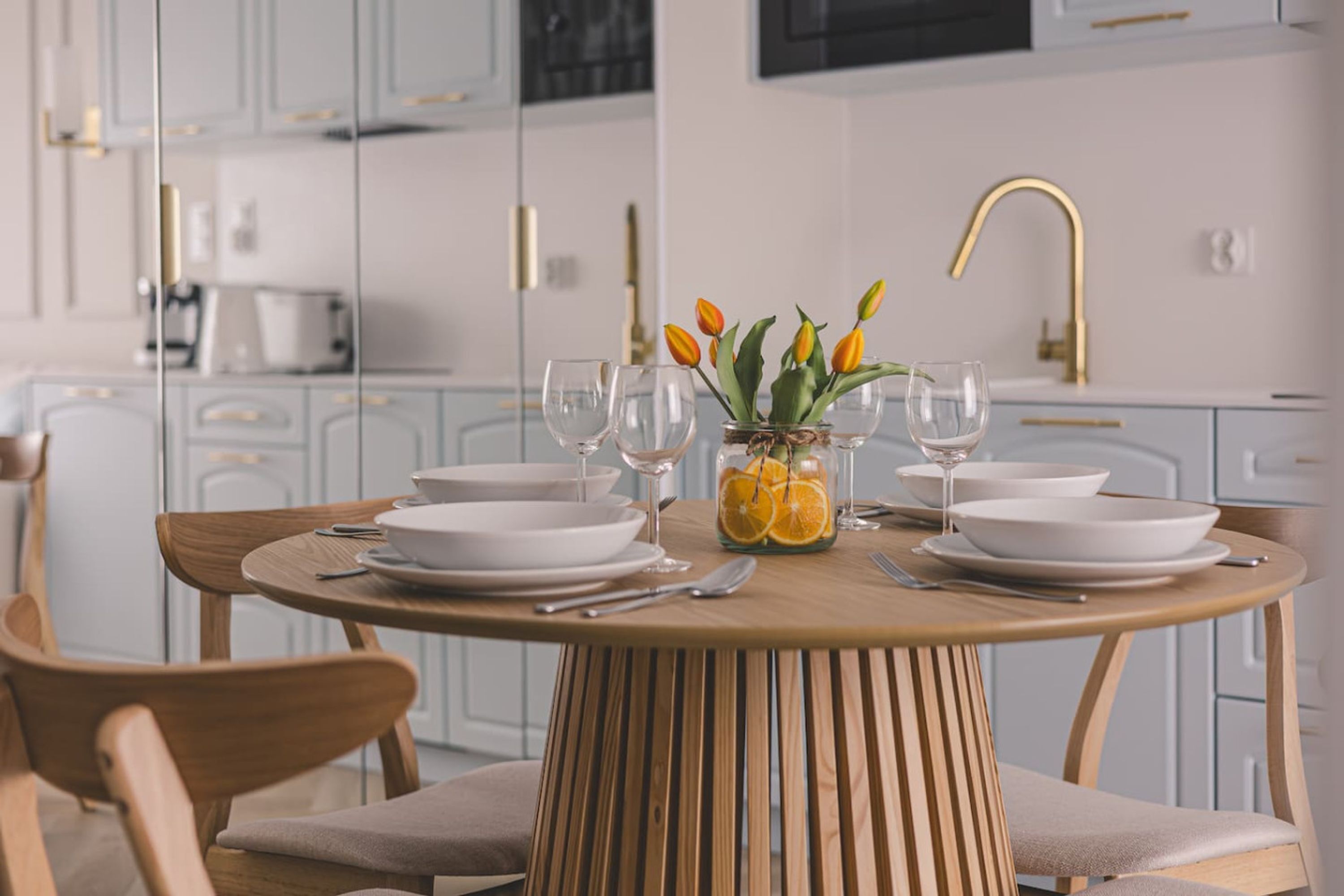 A modern kitchen with white cabinets, a round wooden table set for dining, and a vase with tulips and oranges on the table.