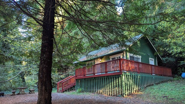 A cabin with a red deck and a green exterior, surrounded by dense forest. - Perfect home base for all things "Redwoods"! Space to park boat. Dog friendly. (Klamath)