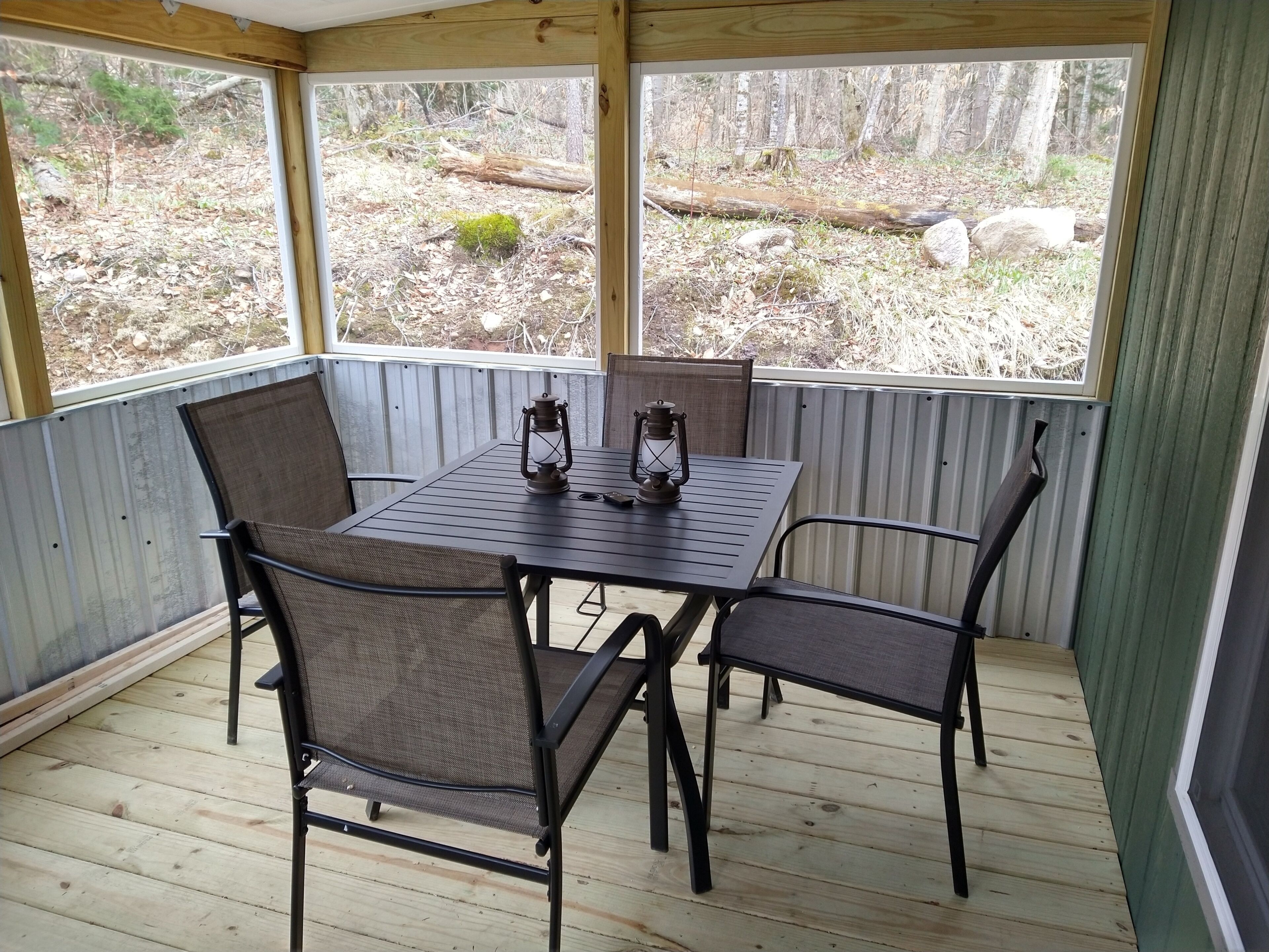 A covered patio with a table and chairs, a lantern, and a view of a wooded area.