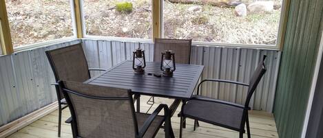 A covered patio with a table and chairs, a lantern, and a view of a wooded area.