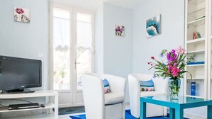 Ein modernes Wohnzimmer mit Fernseher, zwei weißen Sesseln, einem türkisfarbenen Couchtisch und einem blauen Teppich mit Muster.