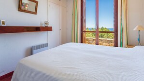 Une chambre avec un grand lit, une étagère en bois, une horloge murale et une fenêtre donnant sur un paysage verdoyant et un plan d’eau.