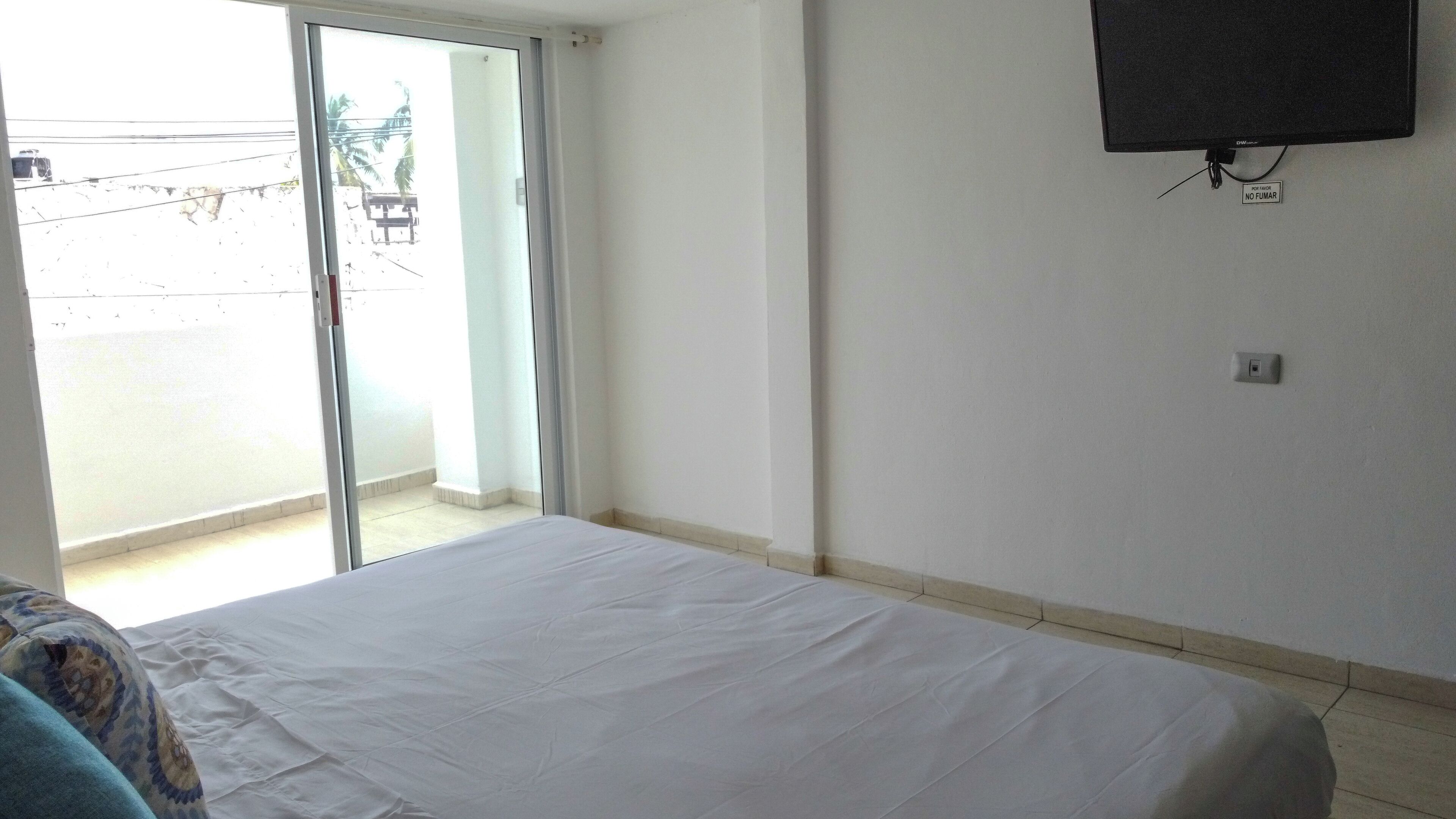 A hotel room with a bed, a TV mounted on the wall, and a sliding glass door leading to a balcony.