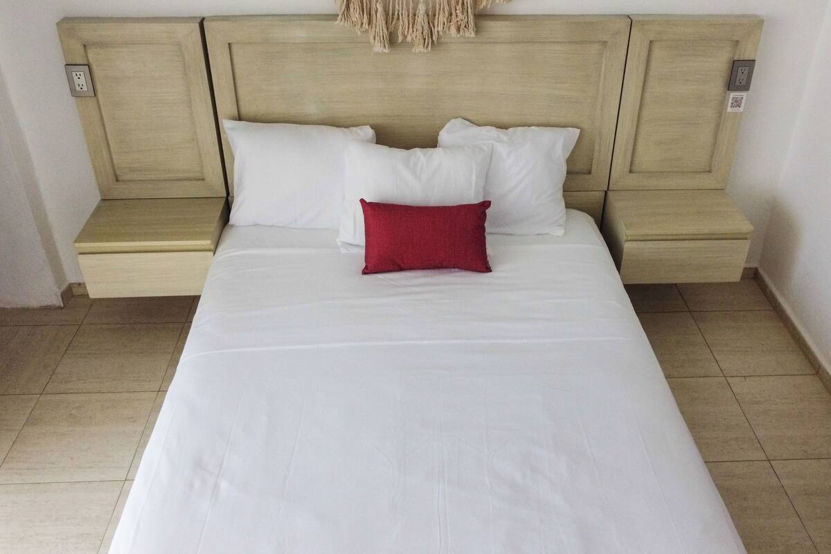 Un letto rifatto con lenzuola bianche e un cuscino decorativo rosso, una testiera in legno e un piccolo comodino.