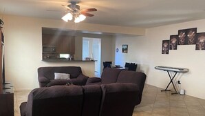 Ein Wohnzimmer mit einem dunkelgrauen Sofa, einem Deckenventilator, einem Flachbildfernseher und einer Küche im Hintergrund.