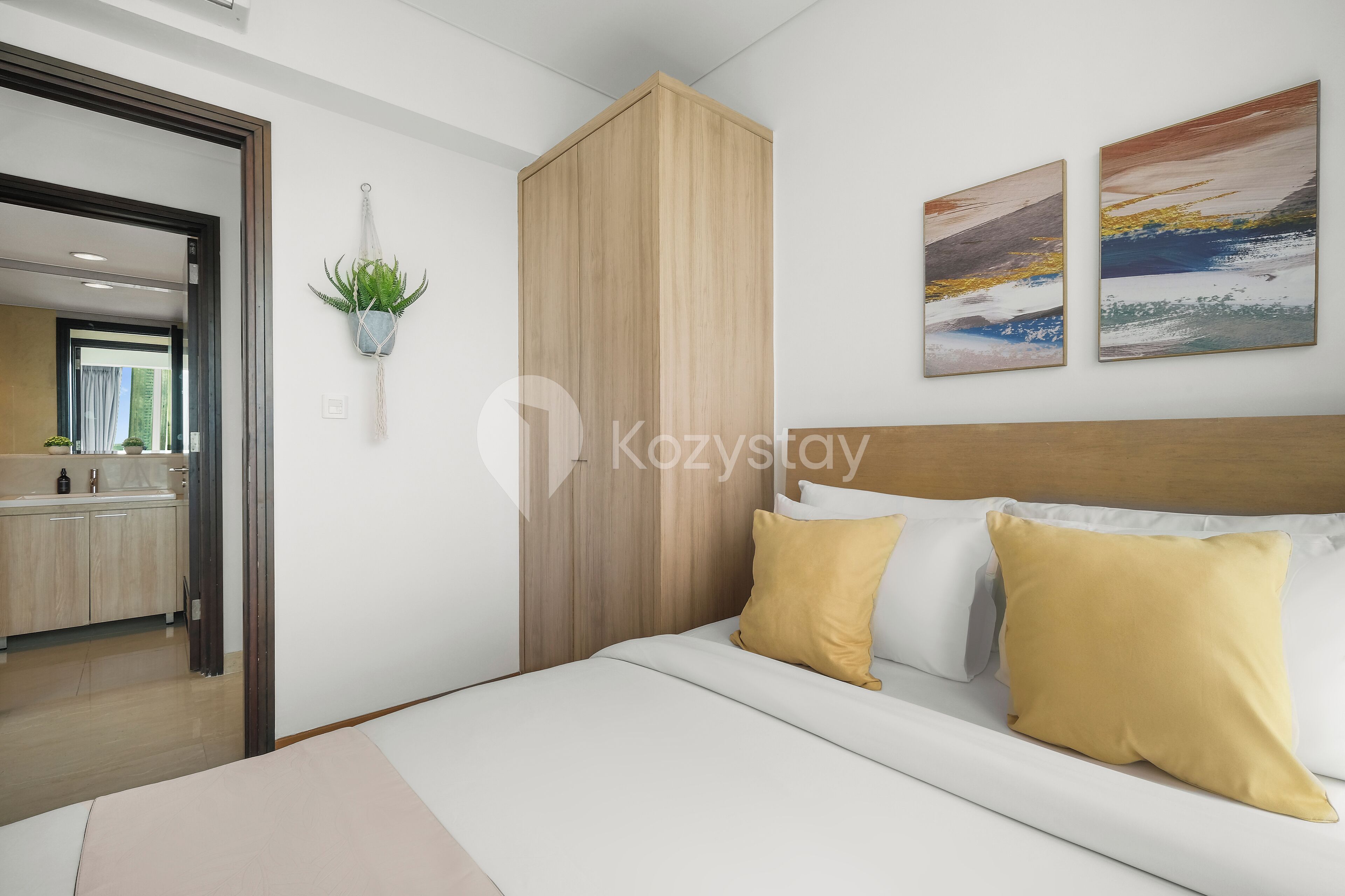 A modern hotel room with a bed, a wooden headboard, a small potted plant, and two abstract paintings on the wall.