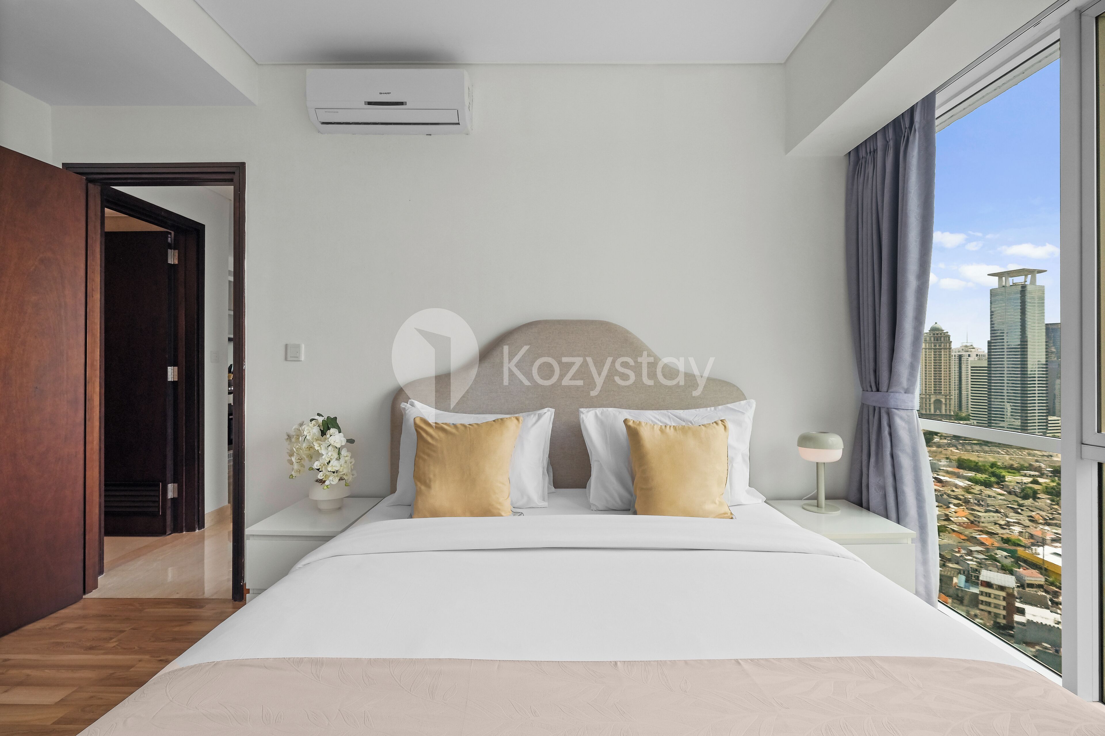 A modern hotel room with a large bed, a nightstand with a lamp, a view of the city through a large window, and an air conditioning unit on the wall.