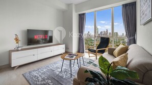 Ein modernes Wohnzimmer mit einem großen Fernseher, der 'NETFLIX' zeigt, einem Sofa, einem Sessel, einem Couchtisch und Blick auf die Stadt durch große Fenster.