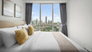 Ein Hotelzimmer mit einem großen Bett, zwei Kissen, einer Kopfteil und Blick auf eine Stadtlandschaft durch große Fenster.