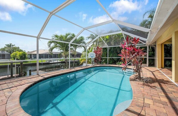 A swimming pool with a glass enclosure, surrounded by palm trees and a brick walkway. - Sand By Me | Heated Pool | Gulf Access & Dock | Near Pelican Sports (Cape Coral)