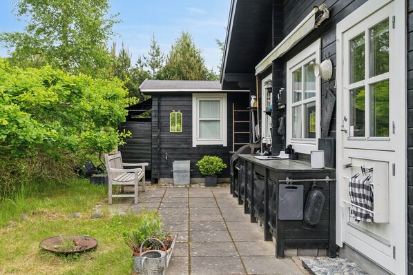 Ein kleines, dunkles Holzhaus mit Terrasse, Bank und kleinem Garten.