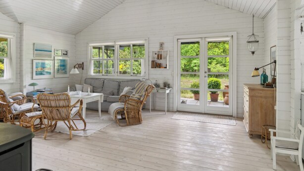 Une pièce spacieuse avec un plafond haut, des murs en bois peints en blanc, une grande fenêtre, un canapé, une table à manger, des chaises en osier et un meuble en bois.