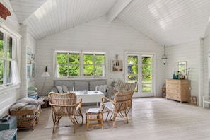 Ein geräumiger Raum mit hoher Decke, großen Fenstern, einer Couch, Korbstühlen, einer Holzkommode und einem Esstisch.
