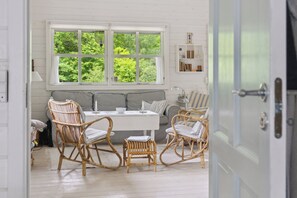 A room with a white wooden floor, a white sofa, a dining table with chairs, and a large window with a view of greenery. - Charming house near private beach and nature (Rørvig)