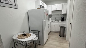 Eine kompakte Küche mit weißen Schränken, einem weißen Kühlschrank, einem kleinen Esstisch mit einem Stuhl und einem Mülleimer.